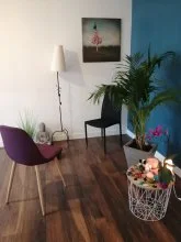 Coin de la salle : chaise violette, chaise noire, lampadaire et tableau, grand palmier devant un mur bleu et petite table fleurie